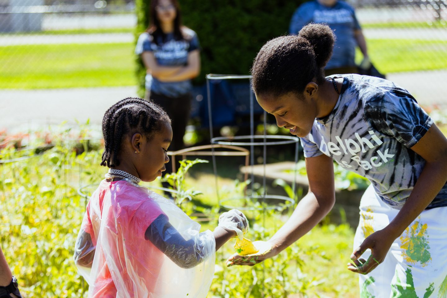 On right, student wearing tie dye t-shirt reading "Adelphi Gives Back" is having their hand painted yellow by a young girl in pink shirt in garden. Student stands in background with arms crossed.