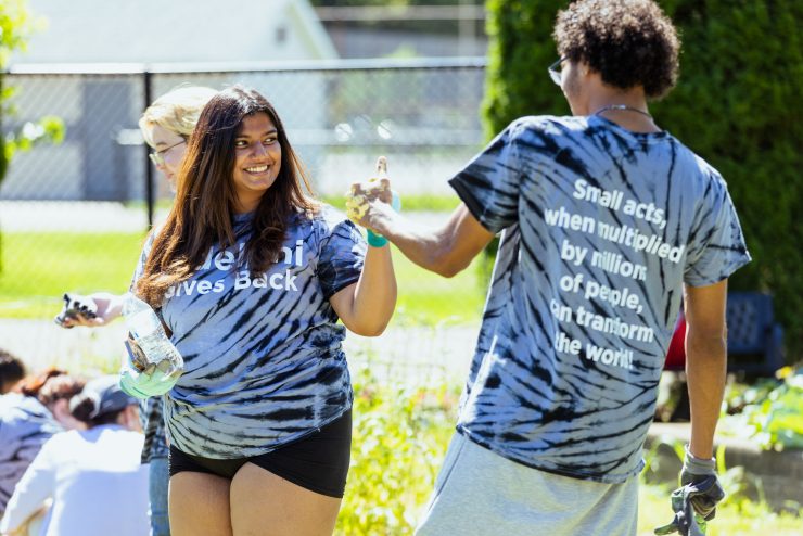 Large group of students wearing tie dye "Adelphi Gives Back" t-shirts hold up certificates. FCAP members on stage behind them.