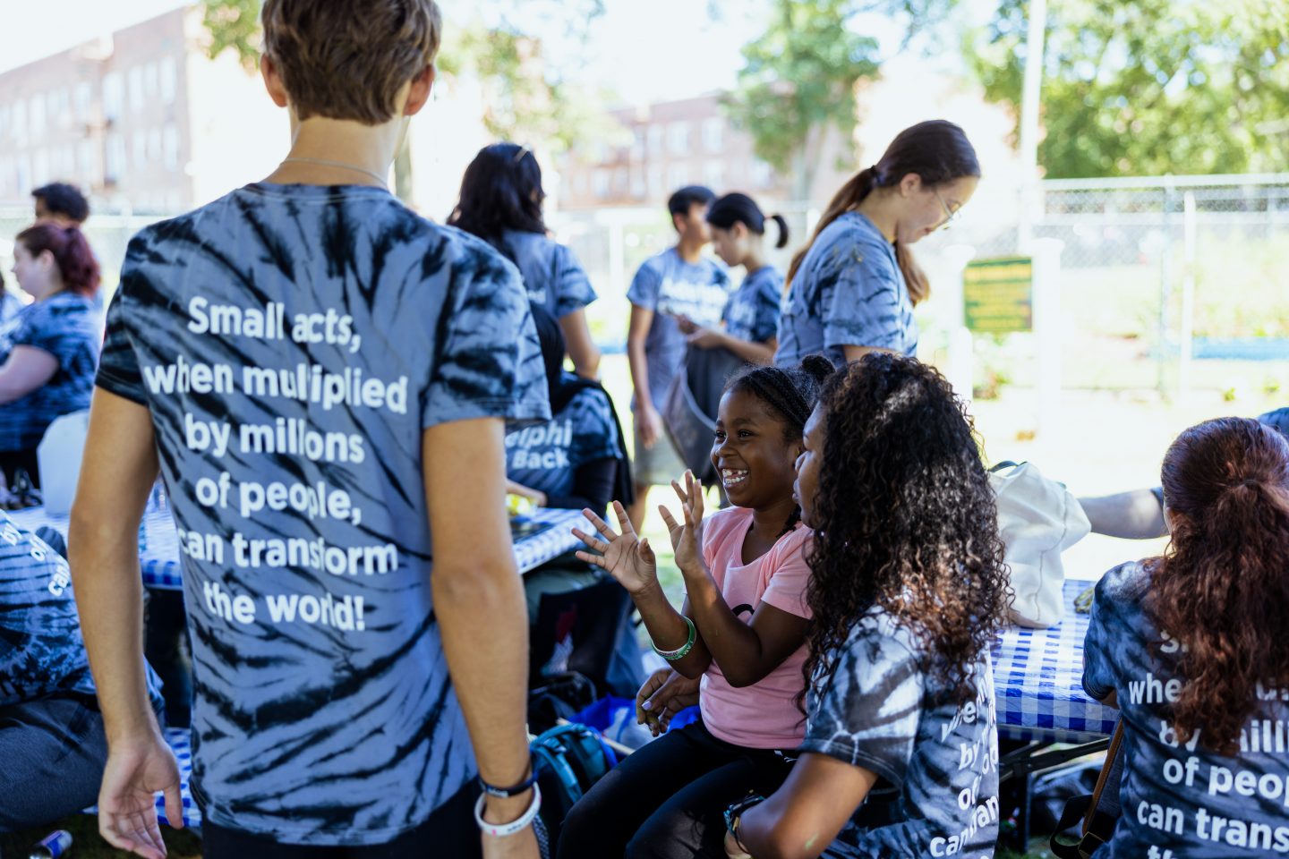 Students all facing away from camera, wearing tie dye t-shirts reading, "Small acts, when multiplied by millions of people, can transform the world!" One student holds child in pink shirt.