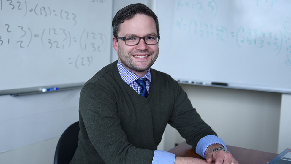 Josh Hiller smiling for the camera, wearing a black v-neck sweater and a blue shirt with a purple tie.