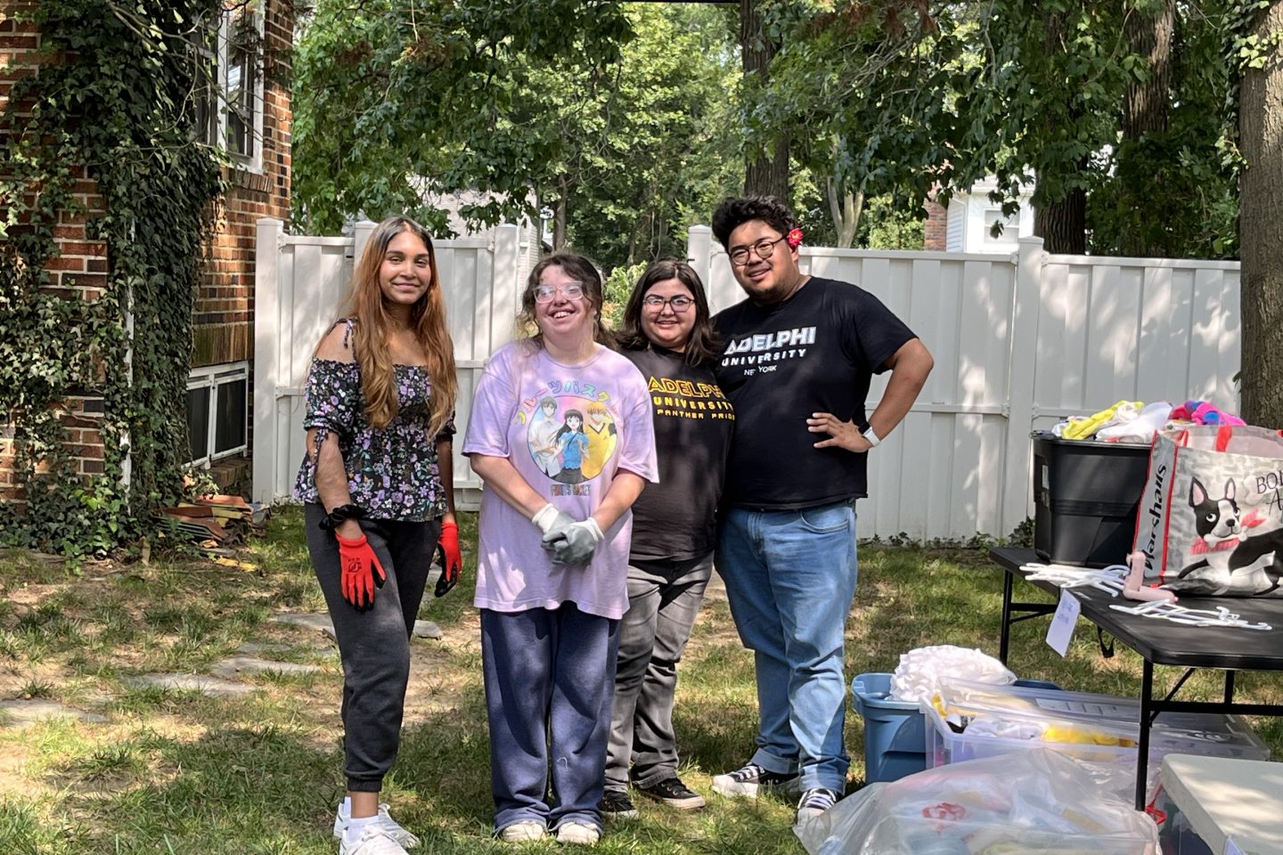 Four students in front of a white fence facing camera, two on left wearing gardening gloves, two on right wearing tie dye T-shirts reading "Adelphi University."