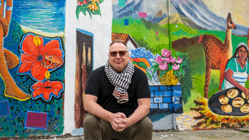 Rob Linné sits on the curb in front of a colorful mural depicting life in El Salvador red hibiscus flowers with a frog one of them, a woman pupusa (the national dish of El Salvador) over an open fire, a llama, a mountain, and a garden wall with a vase of pink tulips on its top.