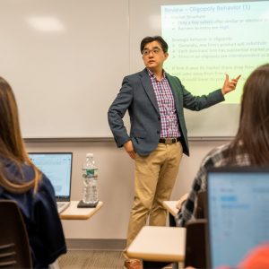 Jason Na stands at the front of his classroom. Two students sitting in their desks, with their laptop computers open, can be seen from behind. Dr. Na is pointing at a slide titled "Oligopoly Behavior" that has been projected onto a screen by an overhead projector.