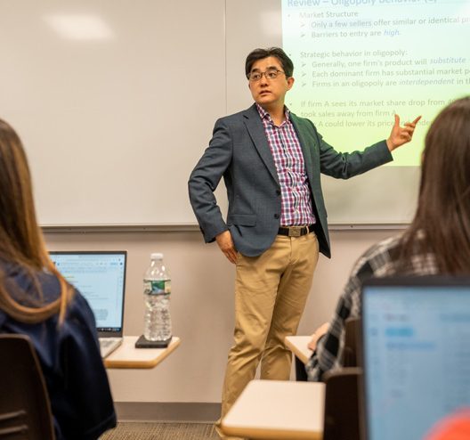Jason Na stands at the front of his classroom. Two students sitting in their desks, with their laptop computers open, can be seen from behind. Dr. Na is pointing at a slide titled "Oligopoly Behavior" that has been projected onto a screen by an overhead projector.