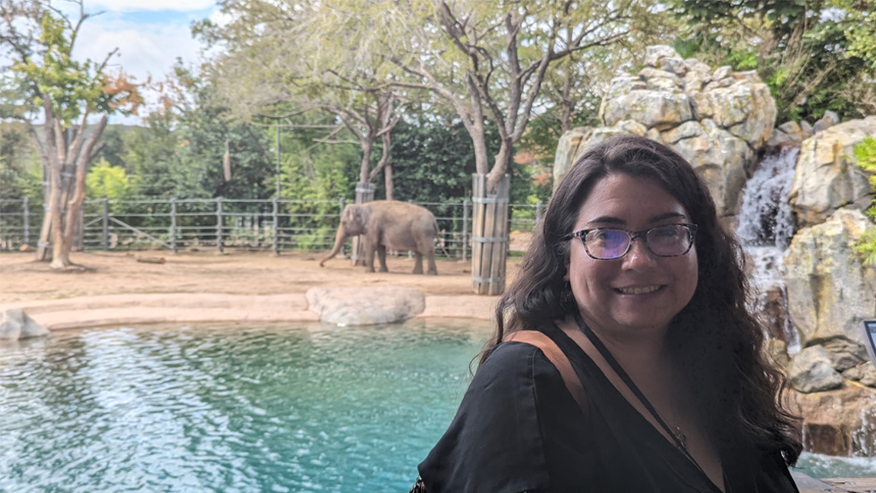A woman wearing glasses in the foreground in front of an elephant in a zoo.