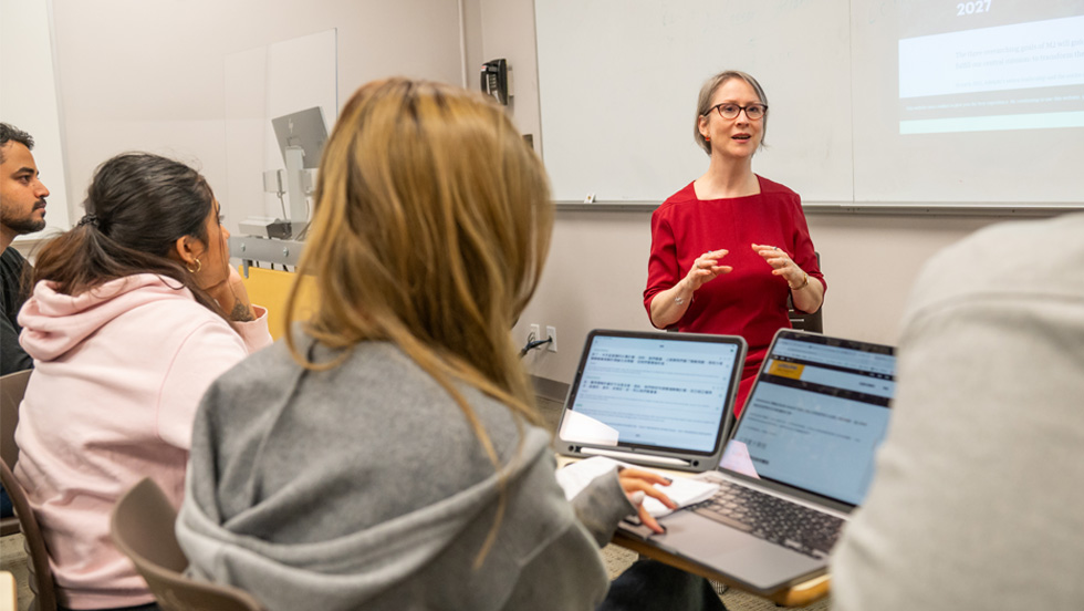 A white female teacher with short, light brown hair and glasses, stands in front of her classroom. Three of her students can be seen at their laptops as they listen to her.