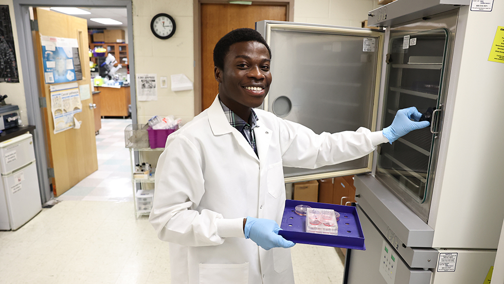 Samuel Sey in a well-lit lab. Wearing a lab coat and blue surgical gloves, he holds a tray with specimens on it, which he prepares to place in a refrigerated cabinet. He is looking at the camera and smiling.