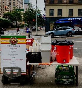 A table set up by a tamal vendor. On it is an orange beverage dispenser, paper cups and a sign that reads Marcellina Tamals