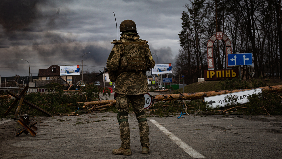  A soldier in camouflage battle fatigues and a helmet stands on a damaged road. He is seen from behind. A tree has fallen across the road, spreading limbs and pine needles across the asphalt. A road sign, in Cyrillic, points the way to Irpin. Billboards can be seen in the distance. Smoke is drifting across a gray sky following shelling by Russian troops.