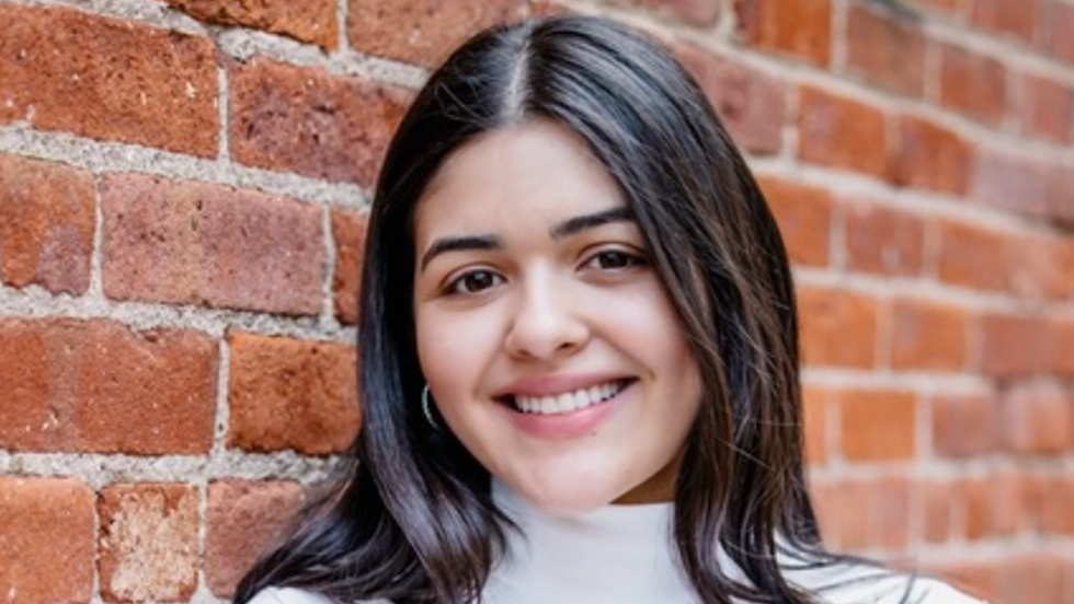 A woman with brown hair and eyes smiling, with arms crossed, wearing white turtleneck