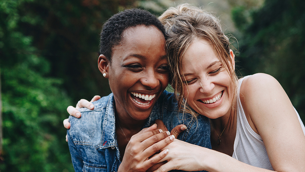 Photo of two women laughing with broad smiles on their face. The women are hugging, with one woman's arm around the other's shoulders. Their heads are touching.