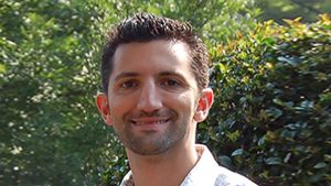 Photo of Dominic Fareri. He is smiling and standing outside, with trees and shrubs behind him.