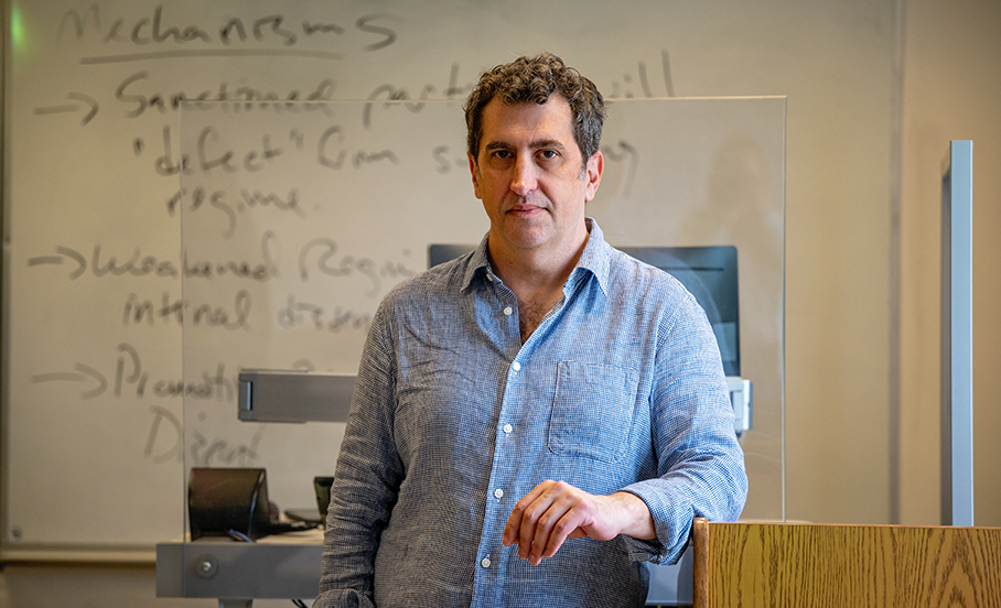 Shawn Kaplan, wearing a blue shirt, standing in front of a classroom whiteboard with notes written on it.