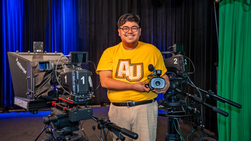 A young man in glasses and a gold shirt that reads AU—for Adelphi University— is smiling, holding a camera. On the left is a camera labeled 1 Autoscript, and on the right, a camera labeled 2.