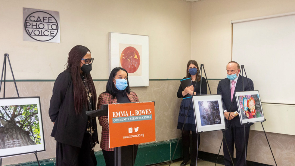 Two students, both wearing medical masks stand at the lecturn while introducing the photovoice project. A sign behind them says "Cafe Photo Voice," while a sign on the lecturn says "Emma L. Bowen Community Services Center" and includes the center's "bowencsc dot org" web address. The students are flanked by three photographs on easels. A woman and a man are at the left looking on.