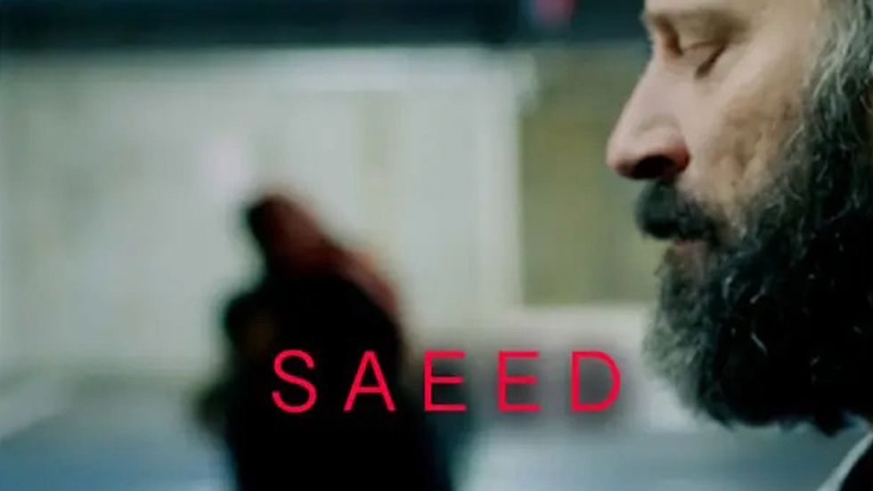 The face of a bearded man, seen from the side, with an out of focus walking past; the name of the film, "Saeed," is superimposed over the photograph.