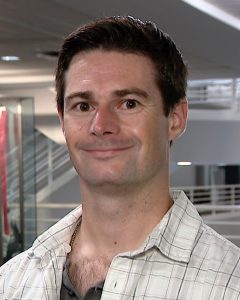 Headshot of a man, body facing slightly away from camera, wearing a plaid button-down shirt. American flag can be seen in the background.