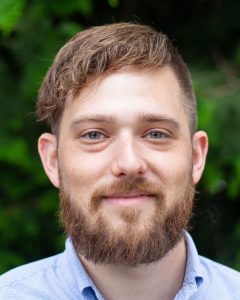 Headshot of man with brown hair and beard against a blurred background of trees