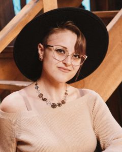 Woman in hat sits on a barrel in front of a wooden staircase.