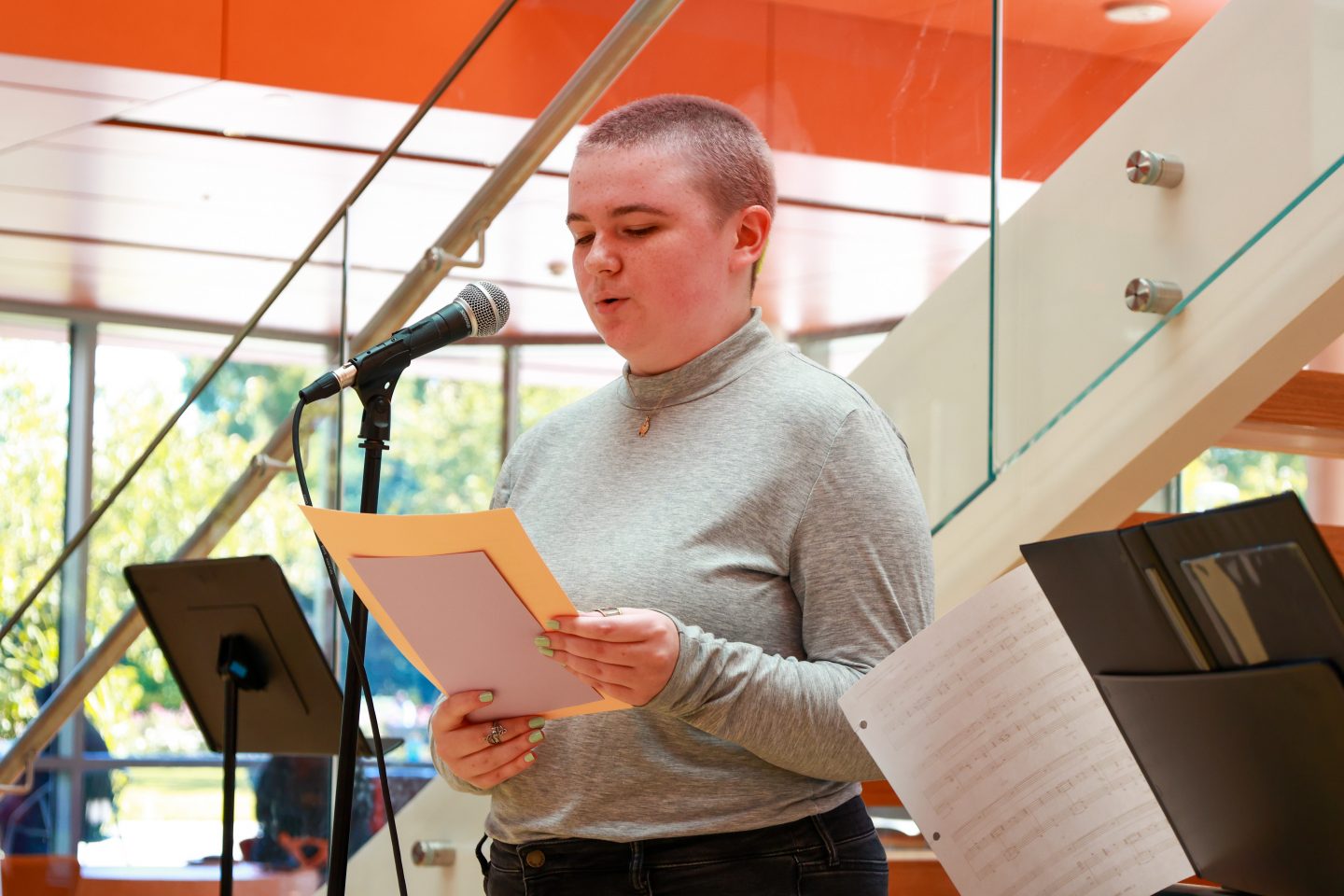 A young woman with a buzz cut wearing a grey shirt is reading from a paper at a microphone
