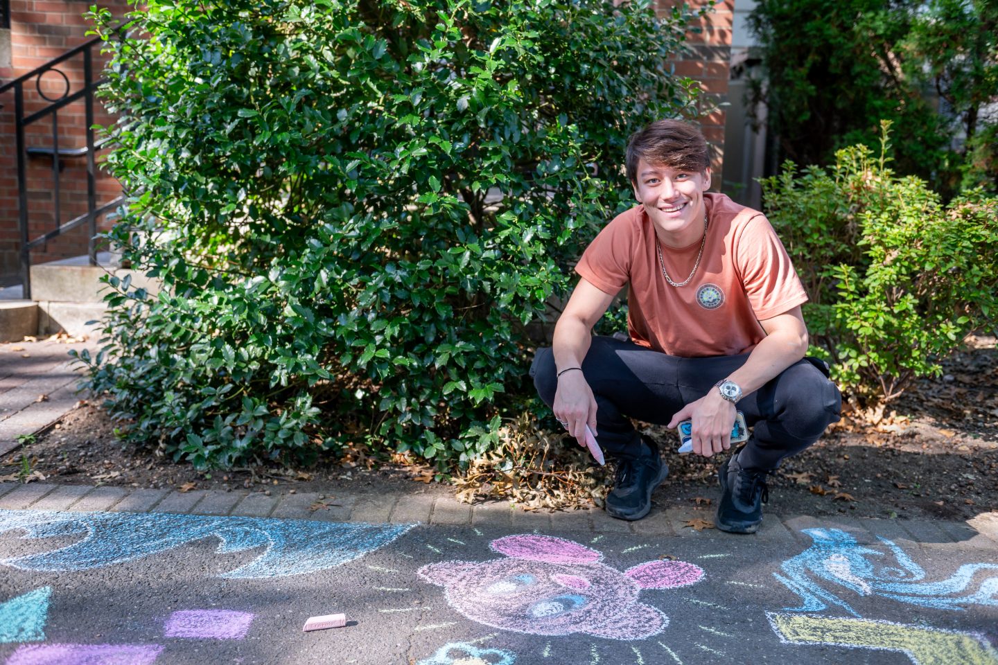 A young man, smiling, squats next to a chalk drawing on a walkway of a happy pink blob.