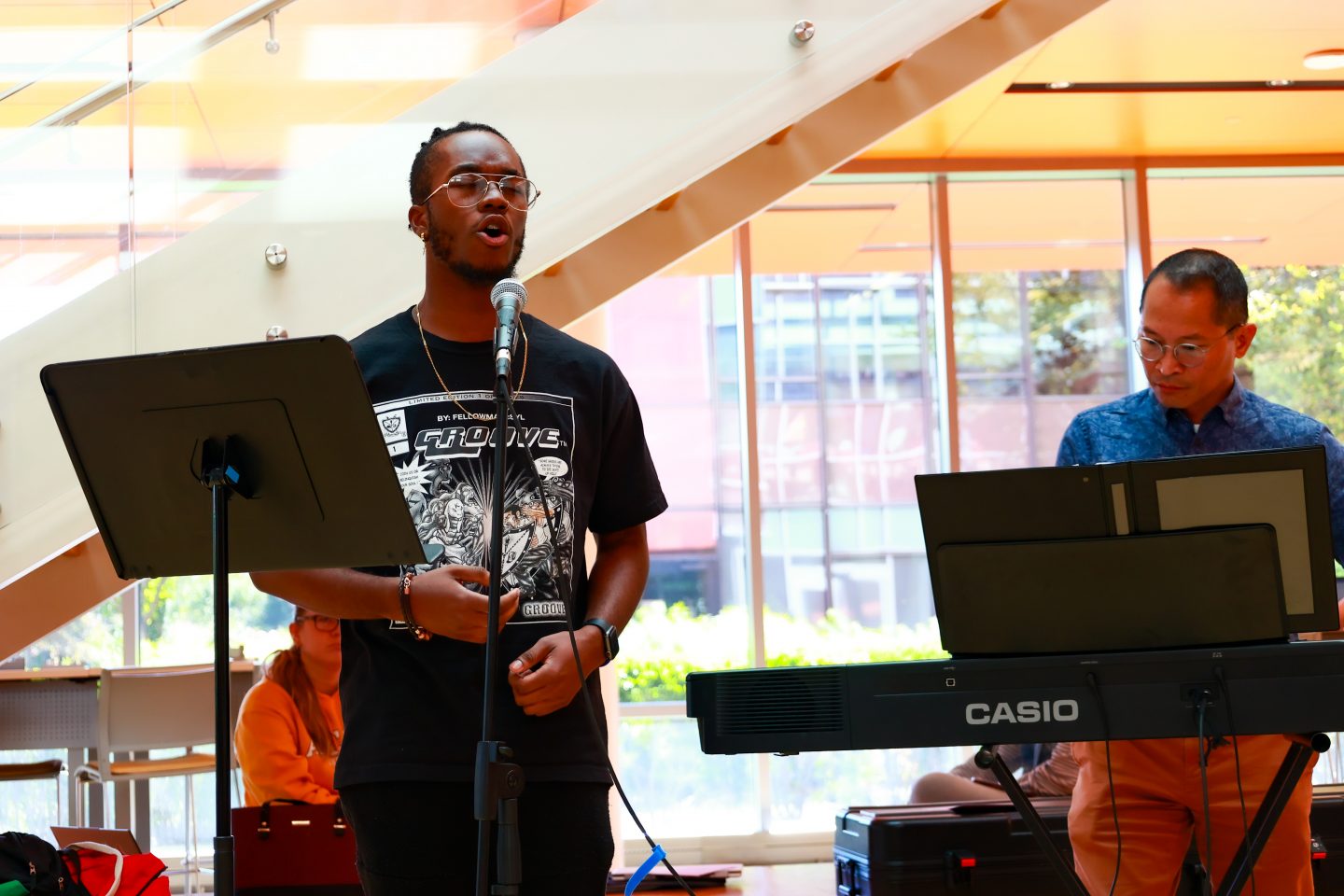 A man wearing a shirt that reads "Groove" is singing. To the right is another man playing a Casio keyboard.