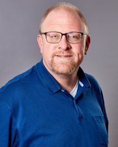 A white man with mustache and beard, wearing glasses and a blue shirt