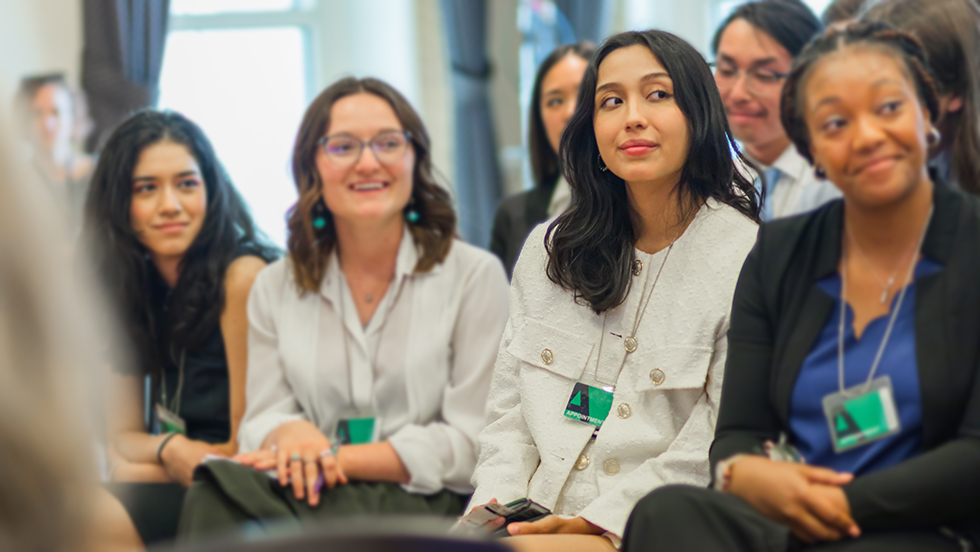 Four young women who are members of the U.S. Digital Corps sit in a row of chairs at a presentation being held in an elegant room at the Eisenhower Executive Office Building, across the street from the White House. All are smiling and wearing government IDs. Three other members of the corps are seated behind them.