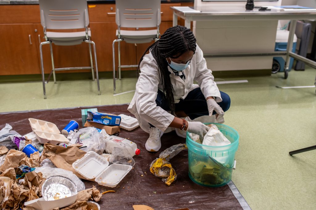 Morenike Olushola-Oni in her lab, separating food waste. She is on the floor, and putting food waste into a bucket while other trash—including styrofoam clamshells trays, aluminum tins, plastic cups and bags used to store food—is heaped on a tarp.