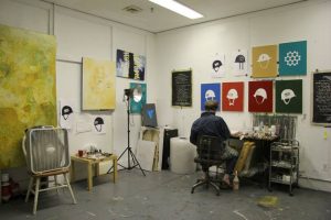 The artist is seen from behind, sitting at a table. We cannot see his face. The white walls are filled with his paintings and drawings, including abstract works and images of military helmets set against flat backgrounds of color. The room is lit by a fluorescent overhead light. There are paint marks are on the gray floor. Art materials sit on a cart and low tables. A roll of bubble wrap and a floor lamp are against the wall, and a whirring box fan sits on an old wooden chair.