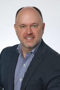 A white man wearing a striped button-down shirt and jacket