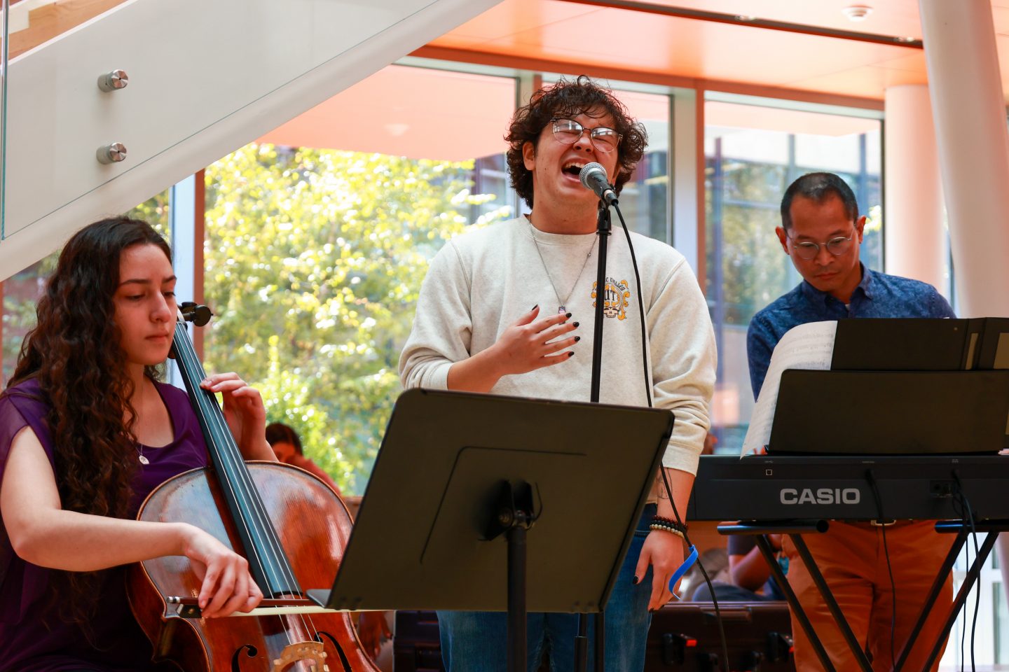 A young man sings at the microphone. To the left is a woman playing the cello. A man playing the keyboard is at right.