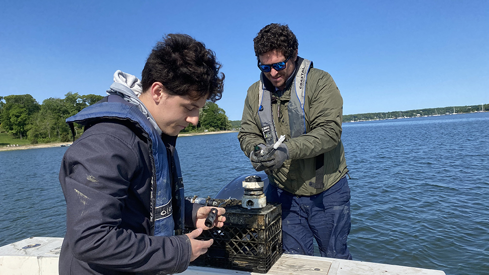 Two men on a boat, holding sensors, with blue water in background under blue sky.