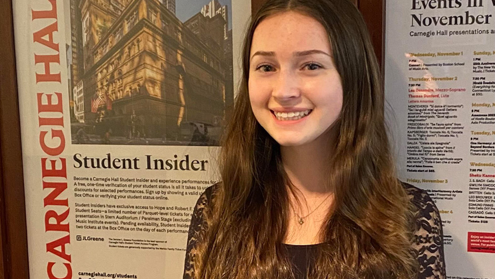 A young white woman with long brown hair, smiling, in front of a poster that reads "Carnegie Hall Student Insider"