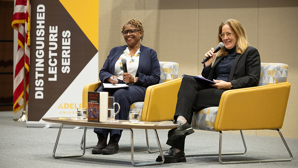 Two women are seated on a stage, looking toward the audience and holding microphones. In front of them is a table with a copy of a book upright. On the left is an American flag and signage that reads: Distinguished Lecture Series, Adelphi University.