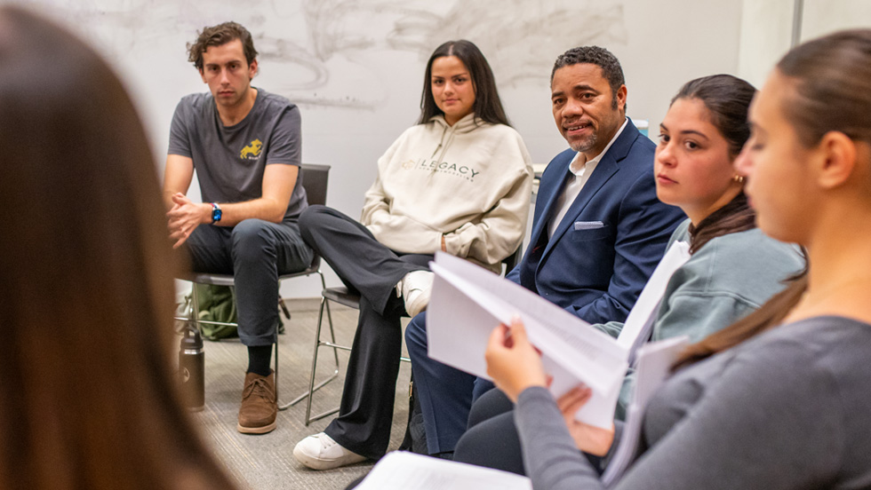 Four students and professor sit in chairs in a rounded formation in a classroom.