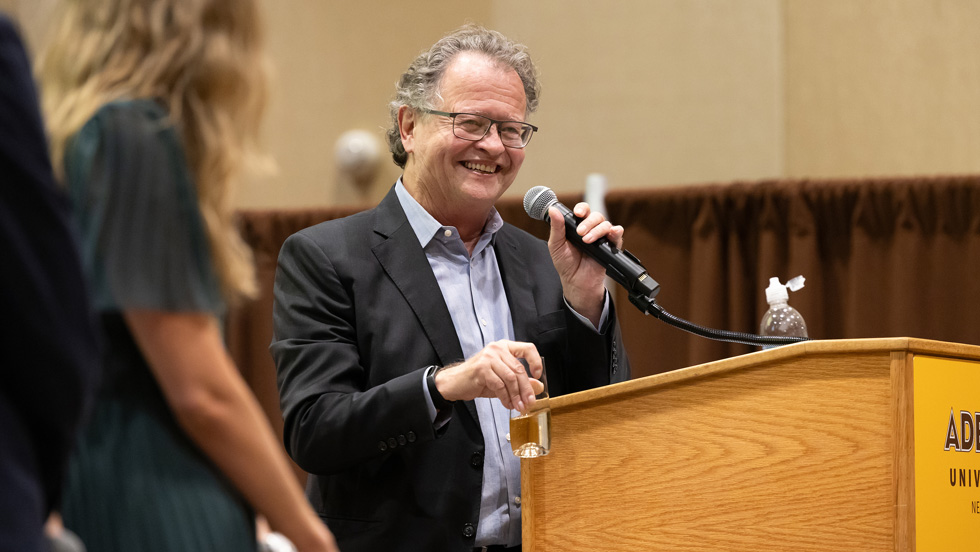 A man, smiling, holds a microphone at a lectern. A woman with her back to the camera stands on the left.