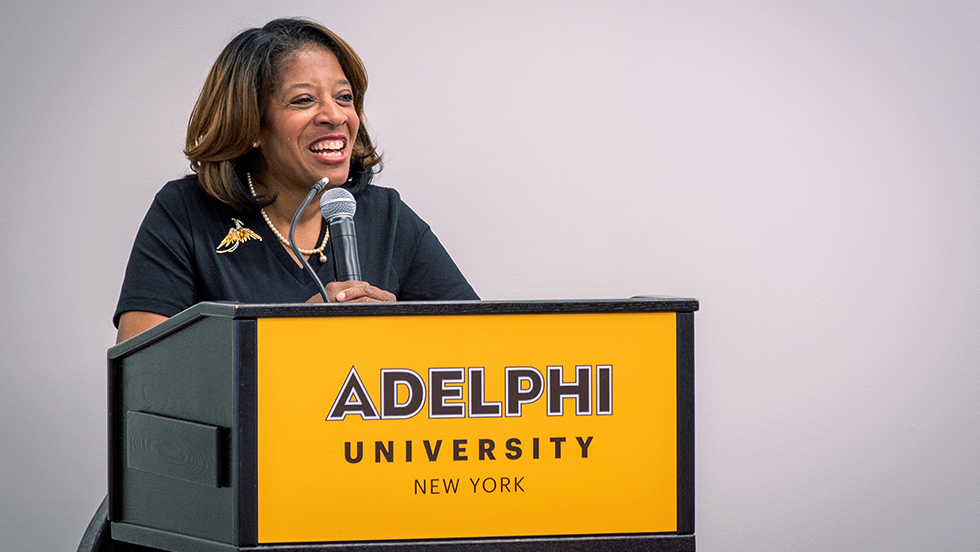 Linda Davis Valdez smiles broadly while giving her address. She is standing before a microphone on a lectern with a gold sign saying Adelphi University, New York.