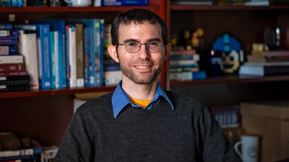 A man with brown hair wearing glasses and a gray sweater, smiling, in front of a bookshelf.