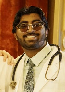 Tommy Joseph smiles for the camera after graduation. He's wearing a lab coat and tie, with a stethoscope around his neck.