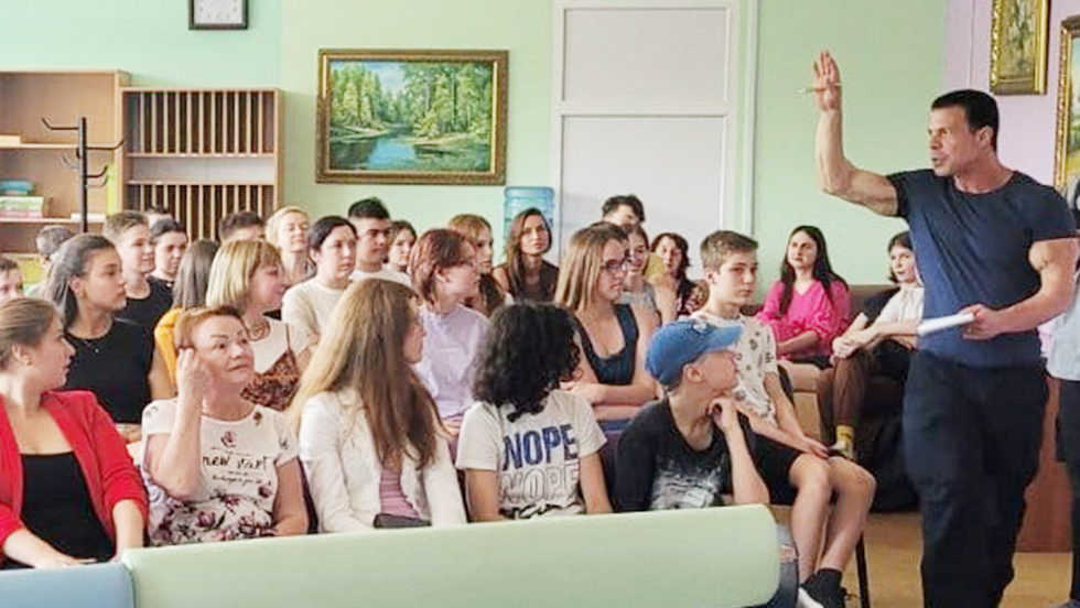 Professor Mirra, holding a book, stands in front of approximately 30 attentive students in a small but bright classroom.