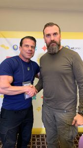 Professor Mirra shakes hands with Oksen Lisovyi, a tall, bearded man wearing military fatigues.