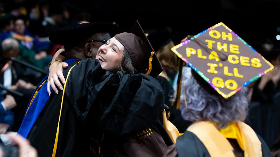 A happy graduate hugs her professor on Commencement day. An older graduate wears a cap that says "Oh, the Places I'll Go."