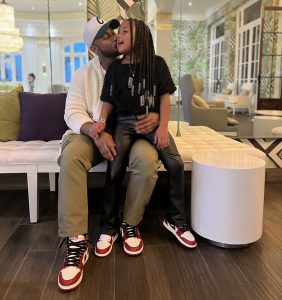 Emma sits on Chad Williams' lap, as he gives her a kiss on the cheeks. The two are wearing matching red and white sneakers and are sitting on a white sofa in front of a mirrored wall.