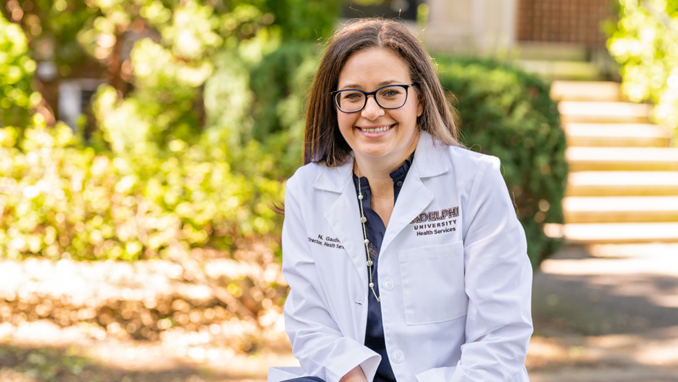 A woman with brown hair and glasses is smiling. She is wearing a white lab coat with the words "Adelphi University Health and Wellness" on the right lapel. Behind her are the grass and trees of the Adelphi campus.