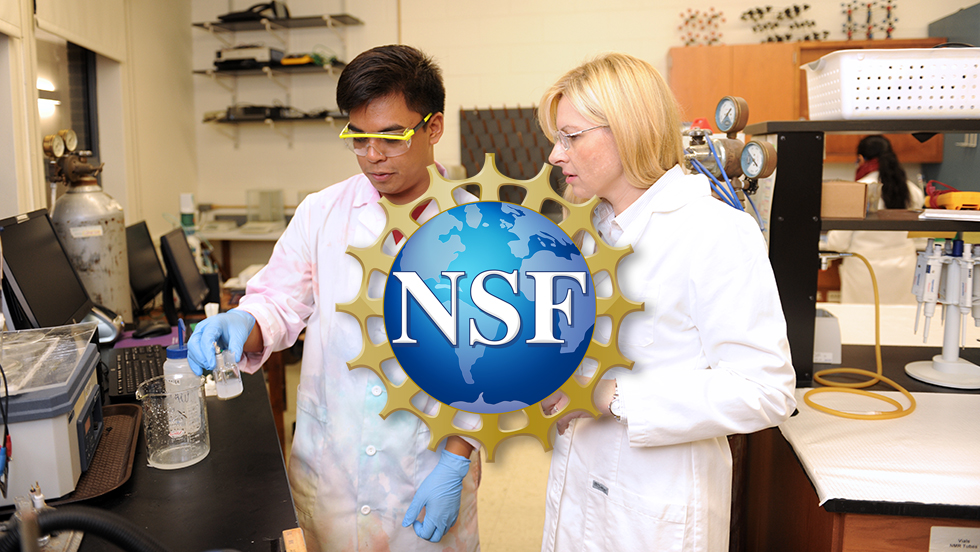 Professor Widera-Kalinowska works with a student conducting an experiment in her lab. Both are wearing lab coats. The student is also wearing protective glasses and gloves and is about to pour a solution into a beaker.