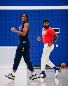 Two Black men in workout clothing are in a gym, doing a standing workout.