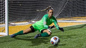 Aliscio, wearing a green goalkeeper's uniform, dives to her left to save a shot that's rolling on the ground.