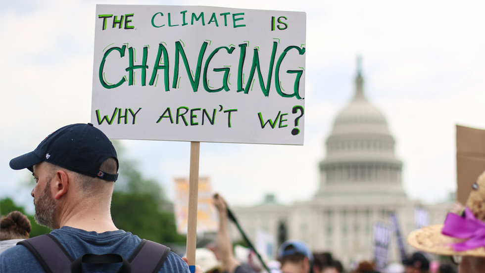 At a protest event in Washington, D.C., with the Capitol Building in the distance, the most prominent is a young man wearing a baseball cap and a backpack, and holding a sign saying "The climate is changing. Why aren't we?"