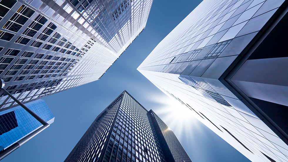 A photo of skyscrapers taken from ground level and looking up at a clear blue sky and a sparkling sun. The photo serves as a visual metaphor for "rising up" to greater opportunity.